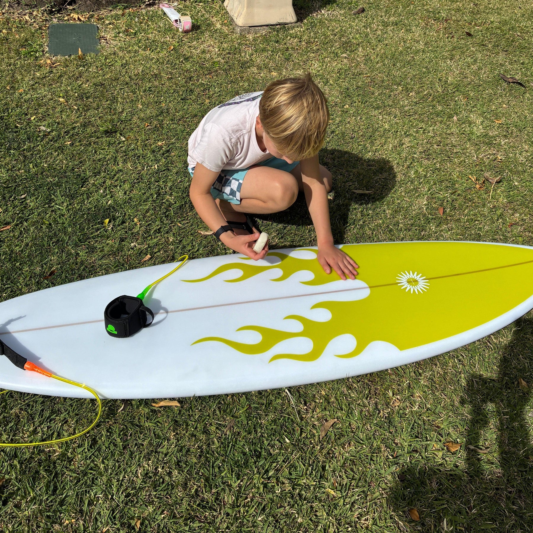 Young surfer waxing a Summer vibes Grom surfboard on grass