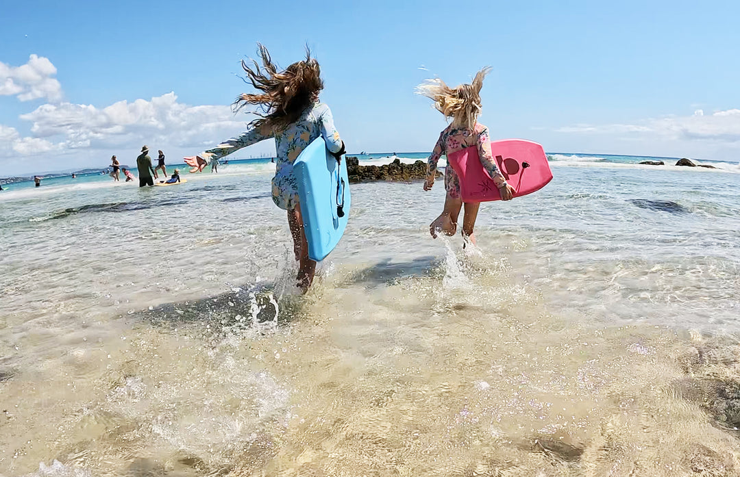 Two children running on a beach with bodyboards, one blue and one pink, under a clear blue sky.
