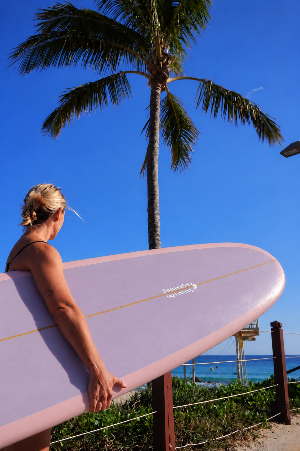 Meghan holding a Summer vibes Classic Noserider surfboard with a soft lilac deck and pink rails at the beach